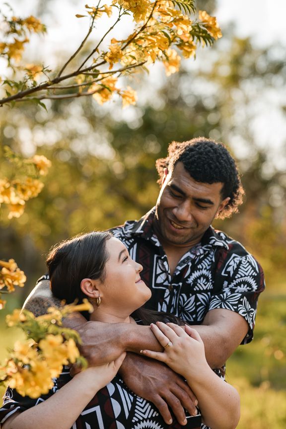 An engaged couple in matching Fijian print shirts embracing under golden blossoms