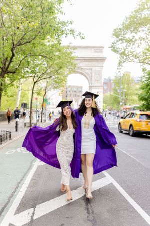 Two graduates in purple academic regalia and caps celebrate on a city street with Washington Square Arch in the background.