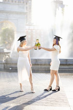 Two graduates in white dresses and caps toast with champagne bottles in front of a fountain on graduation day.