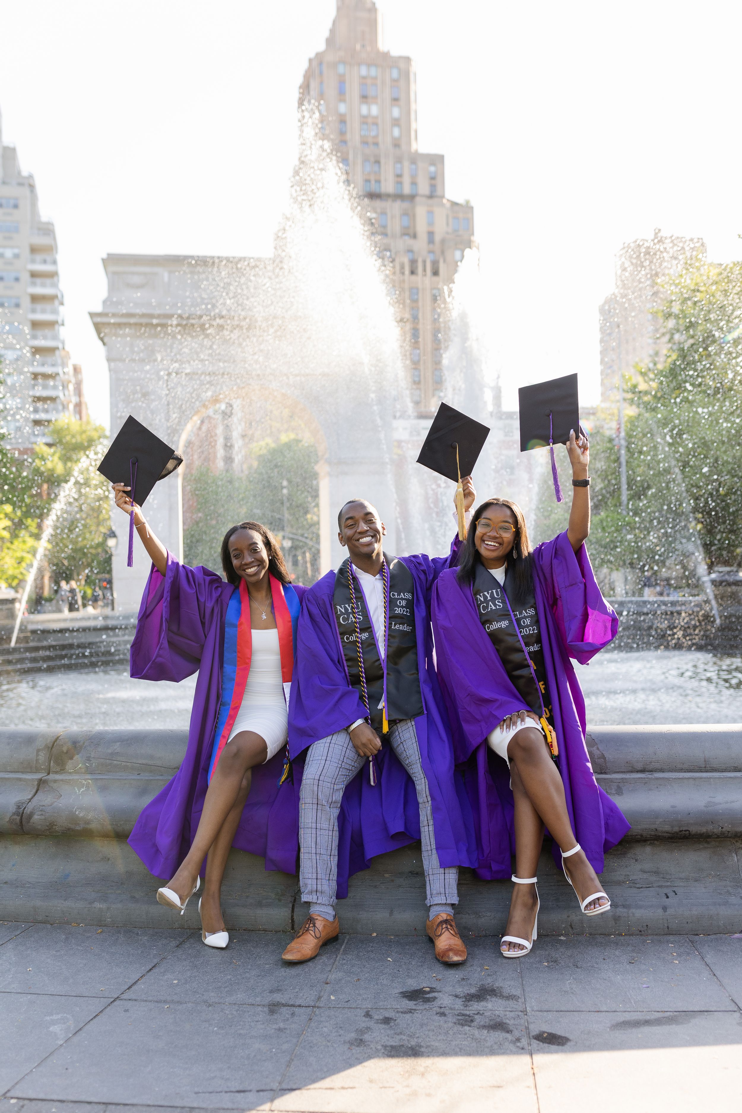 Three graduates in purple robes celebrate with raised graduation caps by a fountain at sunset in an urban setting.