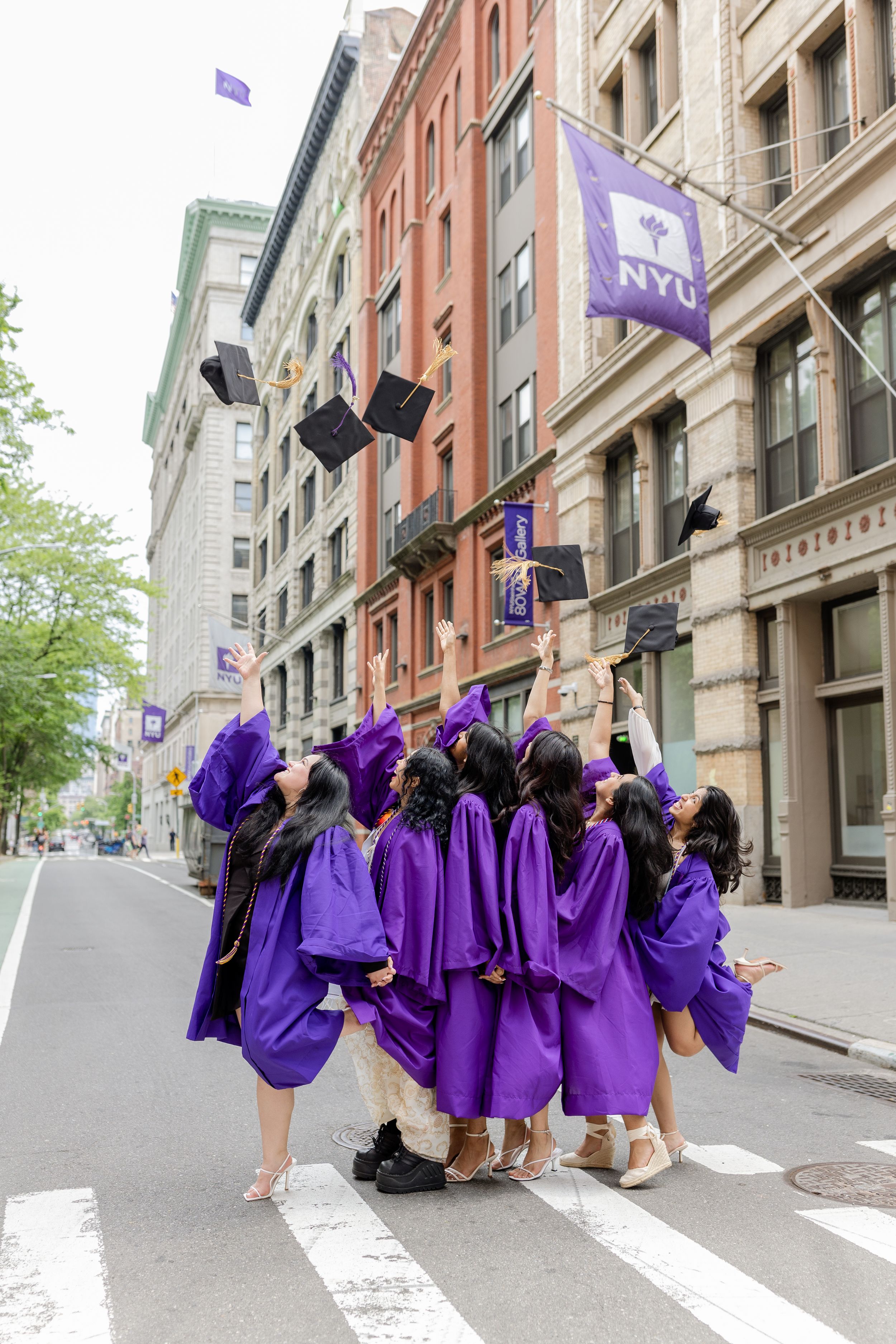A group of graduates in purple caps and gowns walking down a New York City street with NYU flags above.