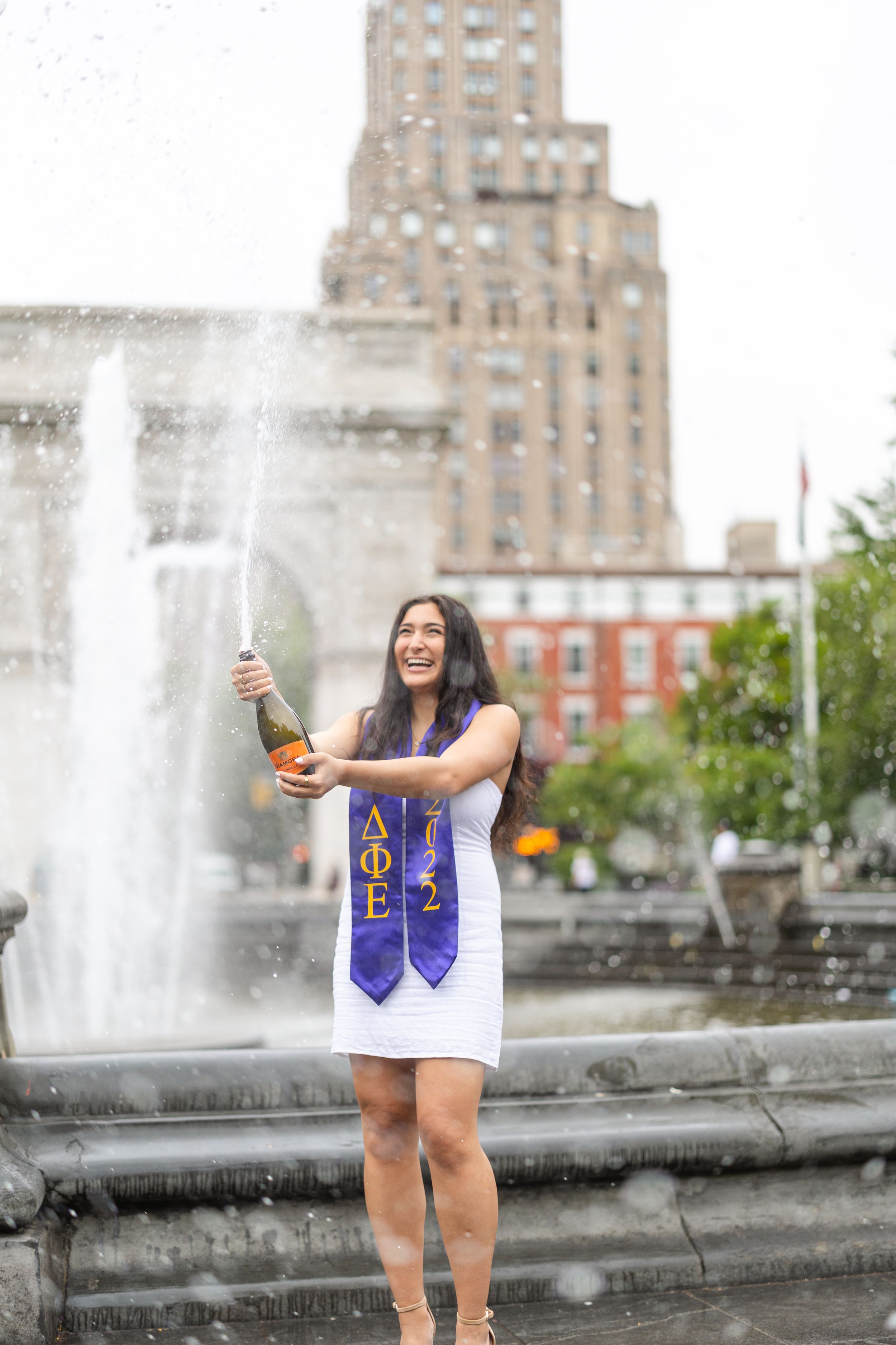 Graduate in white dress with honor cords celebrating by fountain in front of tall city building.