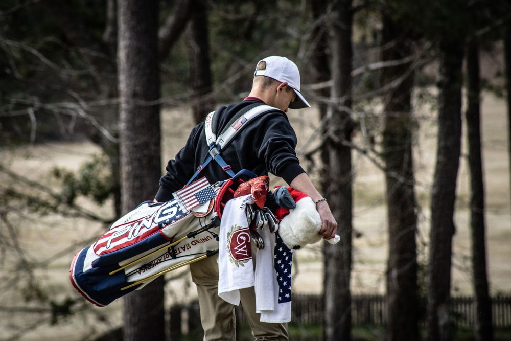 Golfer carrying NC State golf bag through wooded course.