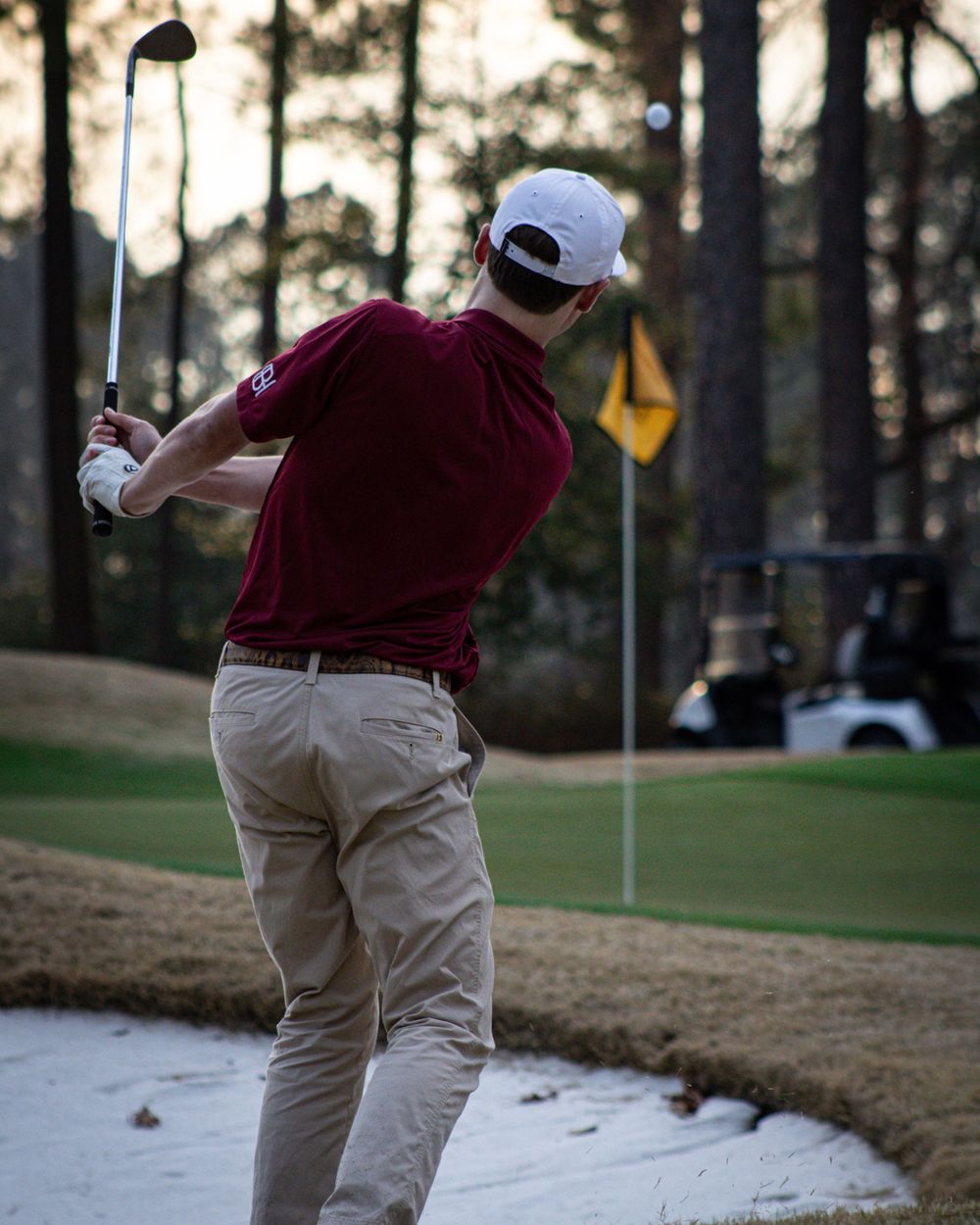 Golfer taking swing at ball from sand bunker during sunset.