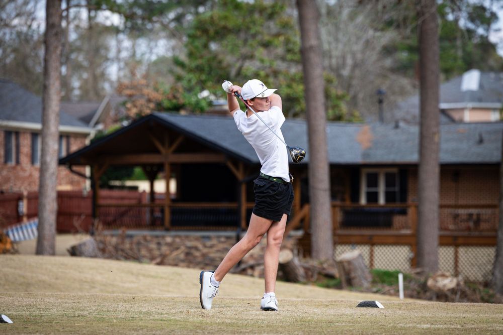 Golfer in white polo shirt and black shorts making a powerful swing on a golf course.