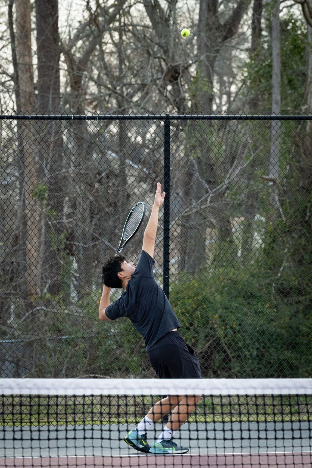 Tennis player executing a powerful serve on an outdoor court surrounded by trees.