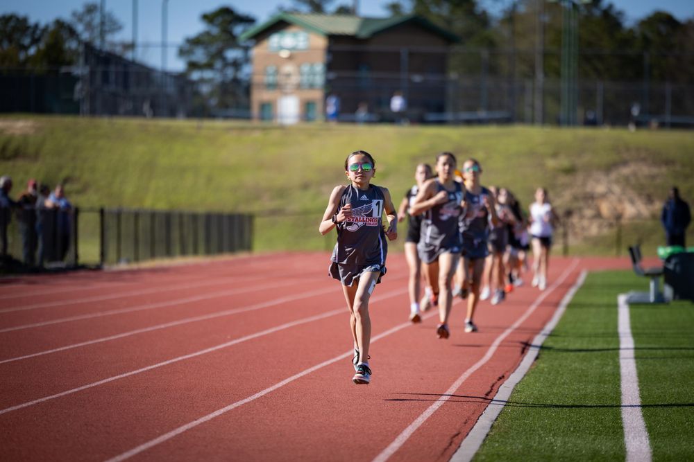 Athletes compete in a track race on a sunny day at an outdoor stadium with spectators watching from the stands.