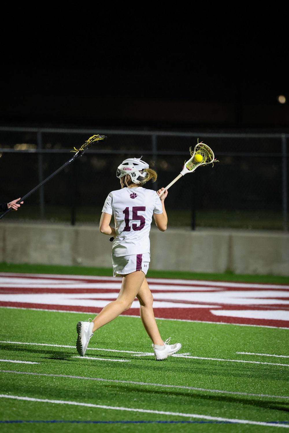 Lacrosse player in white jersey running on a lit sports field at night.