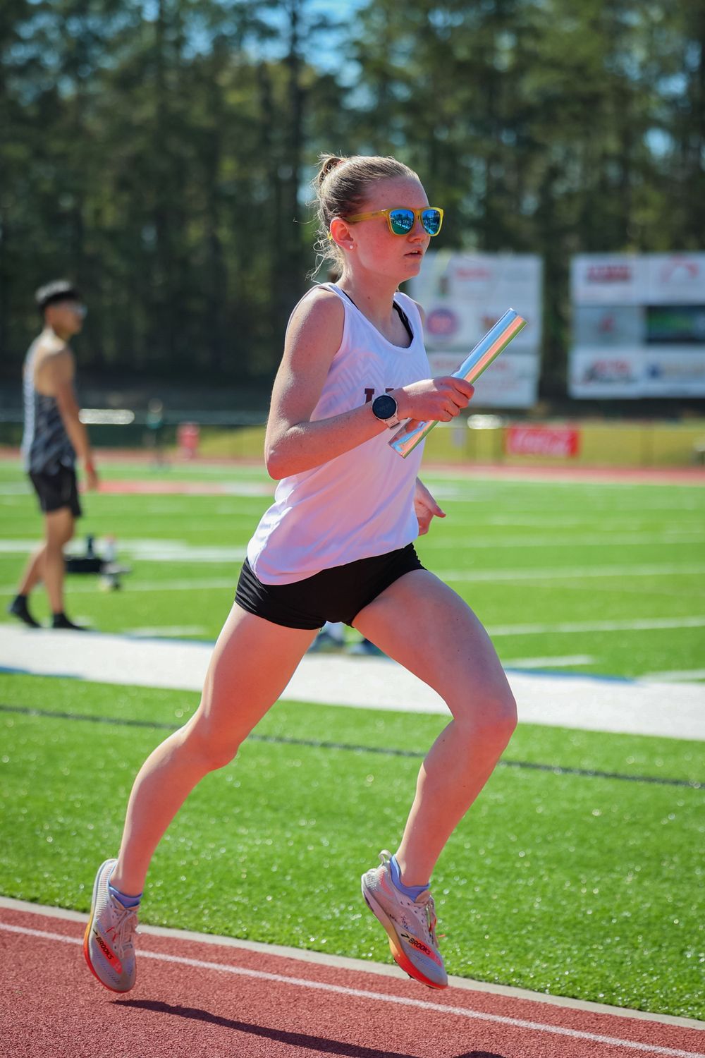 An athlete in sunglasses runs on a red track during a sunny day at a sports competition.