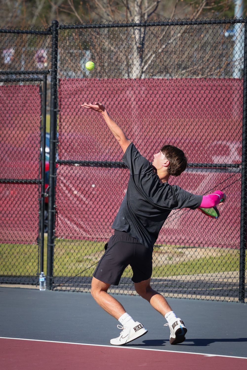 Tennis player serves ball on outdoor court with chain link fence.