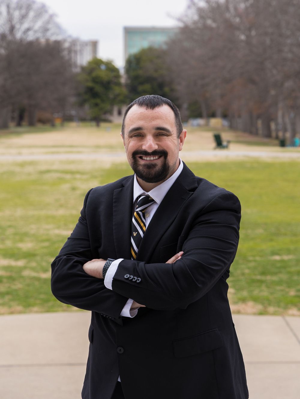 Professional portrait of someone in a dark suit and striped tie standing outdoors in a park setting with arms crossed.