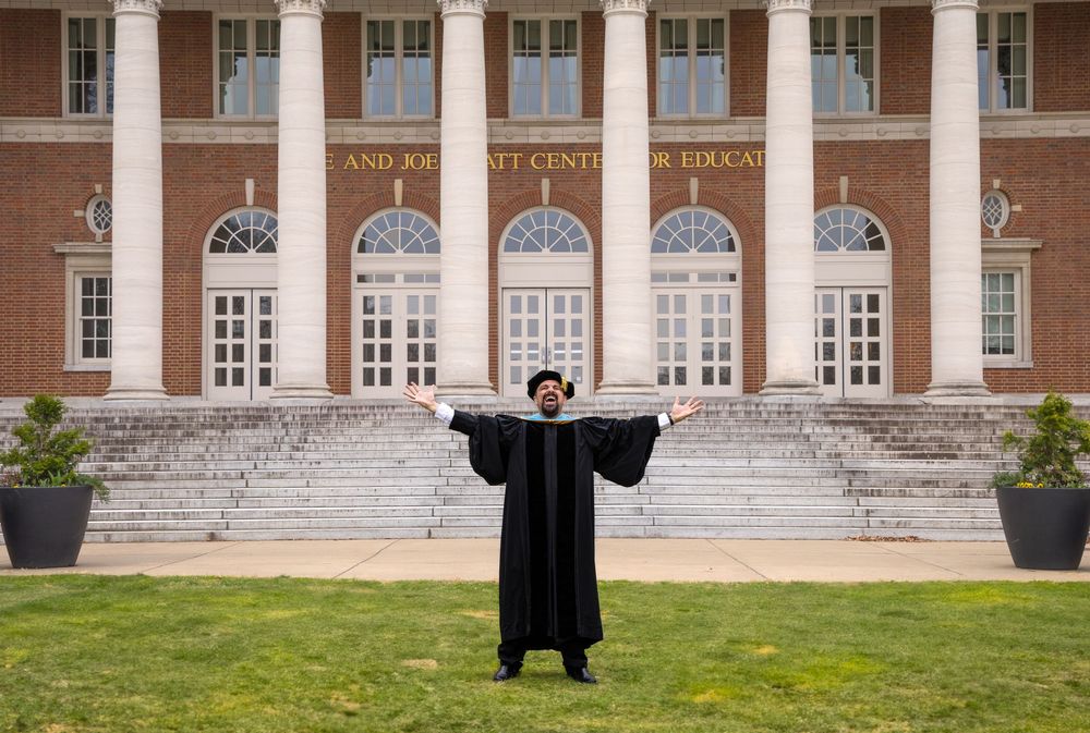 Graduate in black academic regalia stands with outstretched arms on steps of stately brick building with white columns.