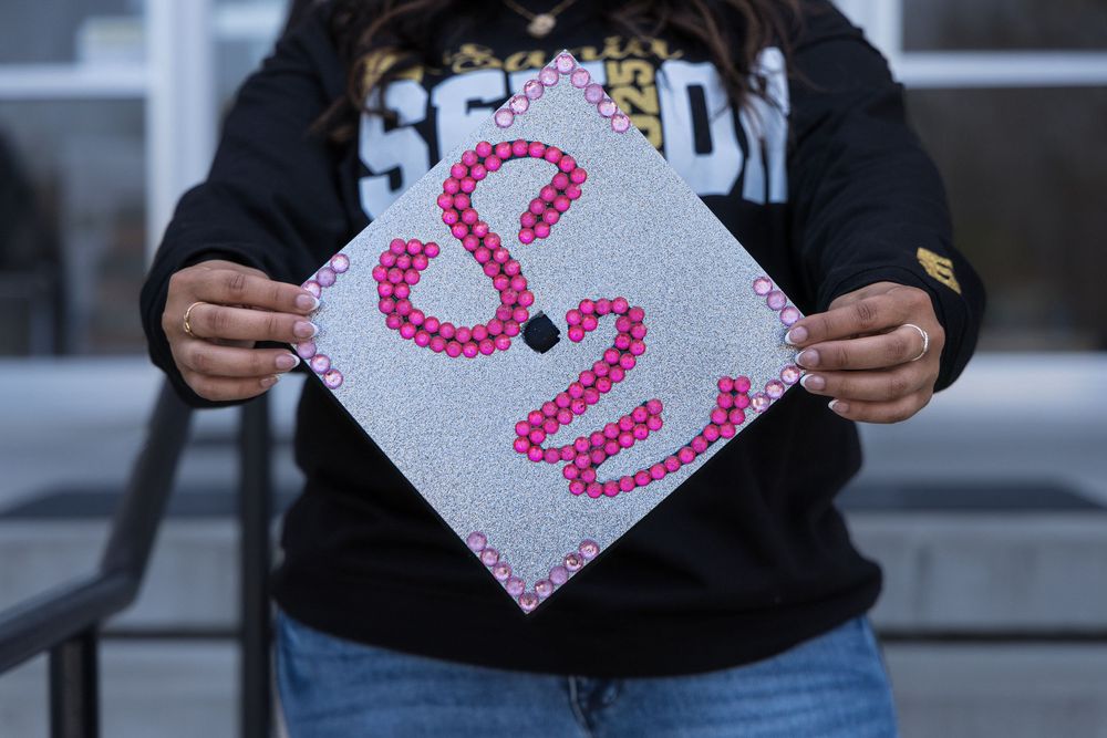A sparkly white graduation cap decorated with pink cursive text is displayed against a dark background.
