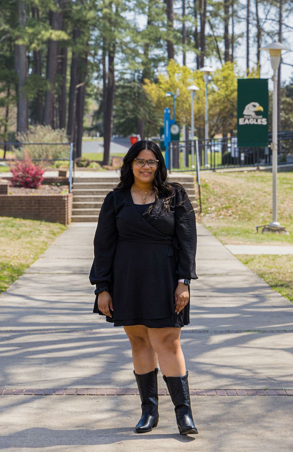 Someone in a black dress and tall boots stands on a brick pathway at a college campus with trees and banners in the background.