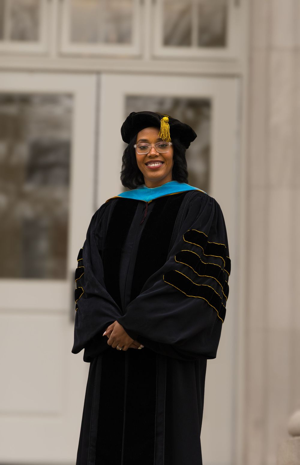 Graduate wearing a doctoral regalia with black gown, light blue hood, and gold tassel at an indoor ceremony.