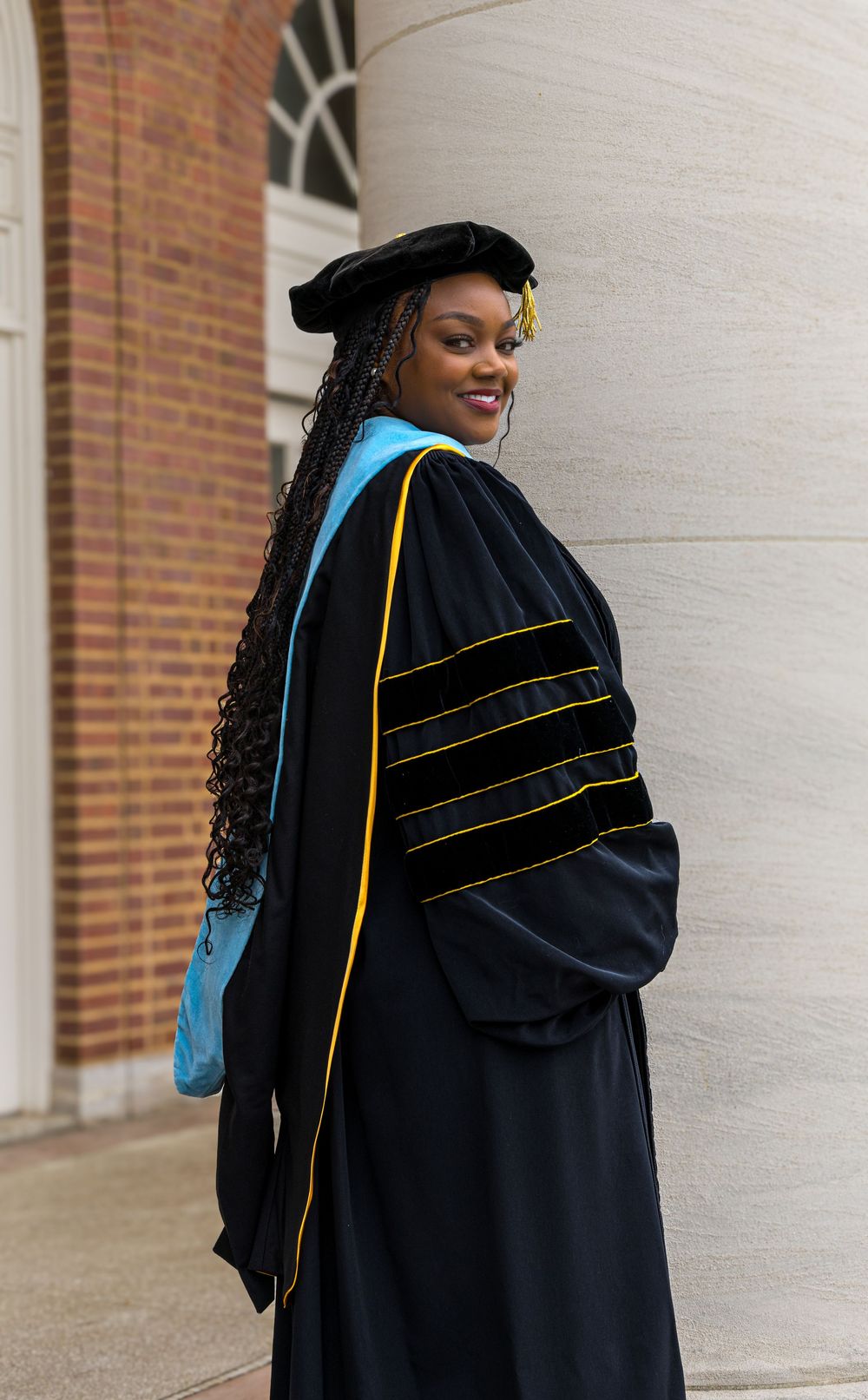 Graduate wearing doctoral academic regalia with blue and gold trim poses in front of brick building.