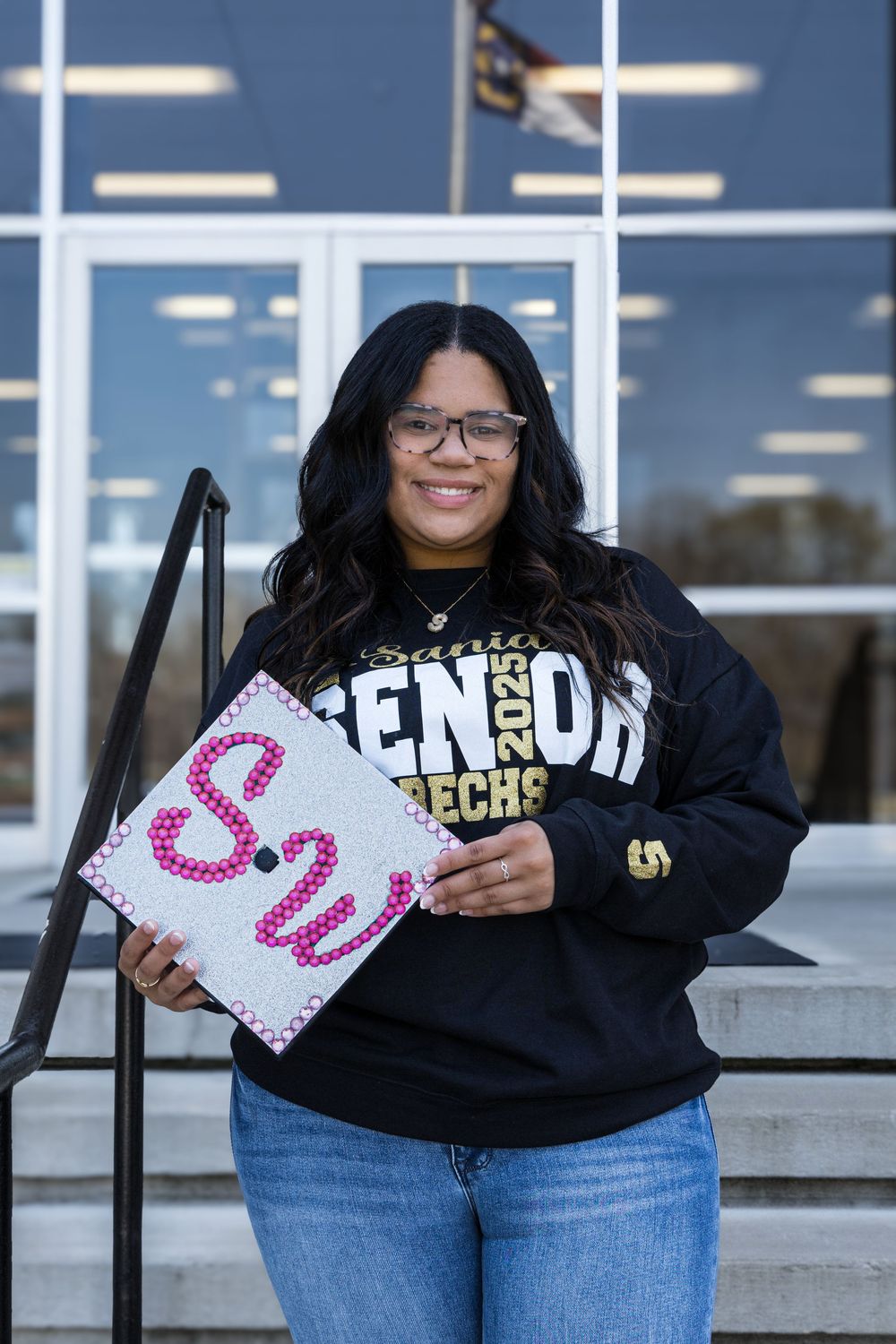 A graduate wearing a Senior Tech sweatshirt holds up a decorated graduation cap with pink glitter letter S outside a building.