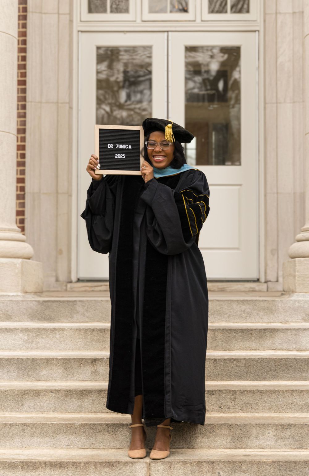 A graduate in academic regalia poses on stone steps while holding up a celebratory sign in front of a columned entrance.