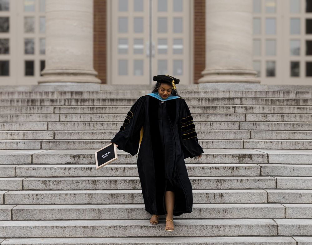 Graduate in traditional black cap and gown celebrates on grand stone steps of university building.