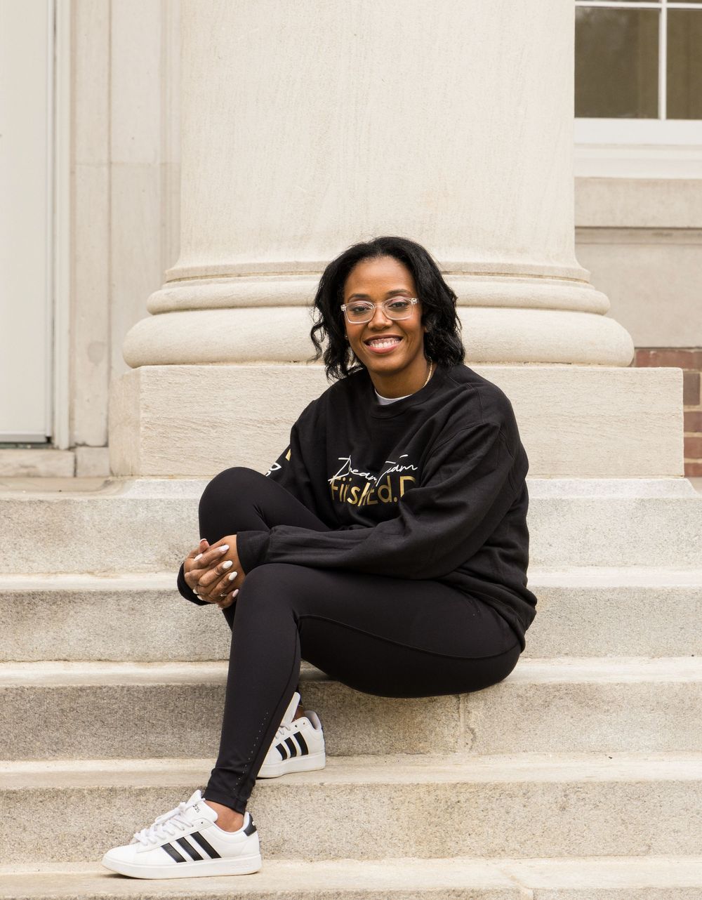 A person in a black sweatshirt and leggings sits relaxed on concrete steps, wearing Adidas sneakers.