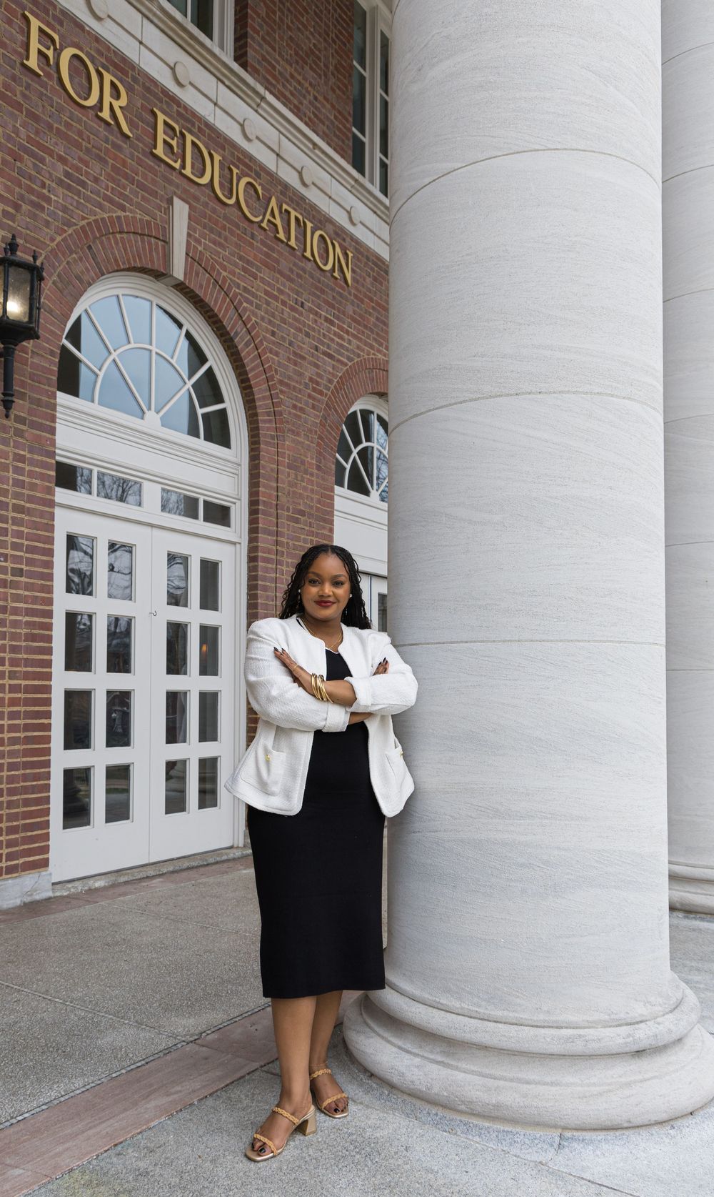 A person stands confidently in front of a brick education building entrance with white columns wearing business attire.