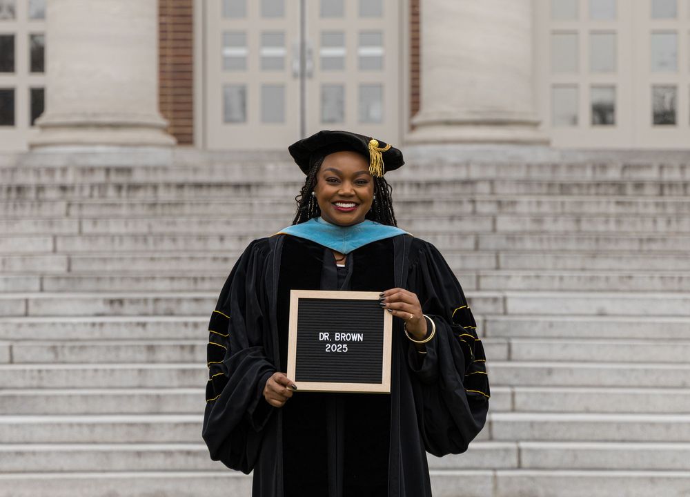 Graduate in traditional black cap and gown holds a small chalkboard sign while standing on steps of academic building.