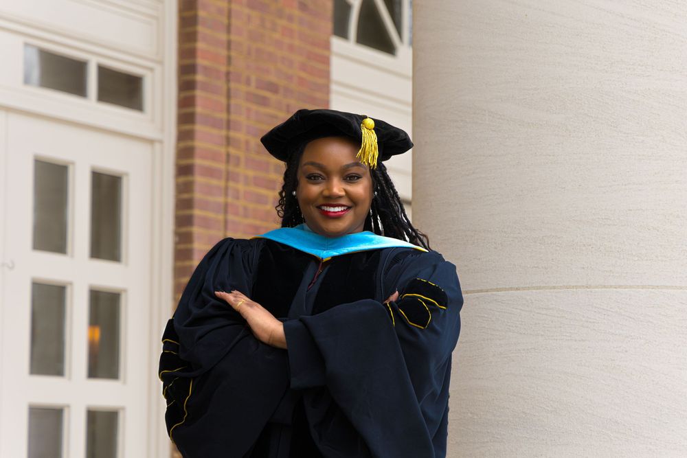 Graduate wearing traditional black doctoral gown and graduation cap with blue accent stands smiling beside white column.