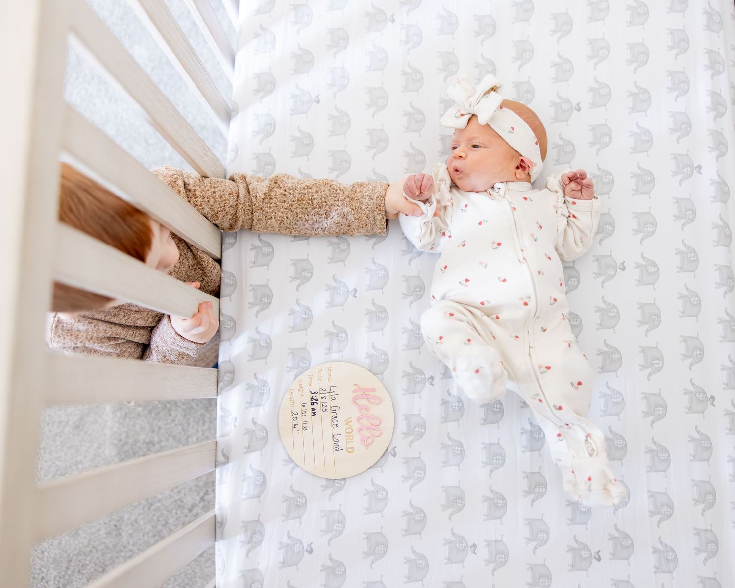 A sleeping baby wearing white lies in a crib with gray elephant patterned wallpaper in the background.