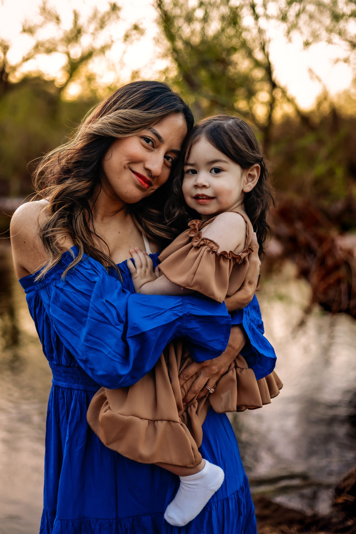 A mother in a royal blue dress and child in brown dress pose together at sunset in a natural outdoor setting.