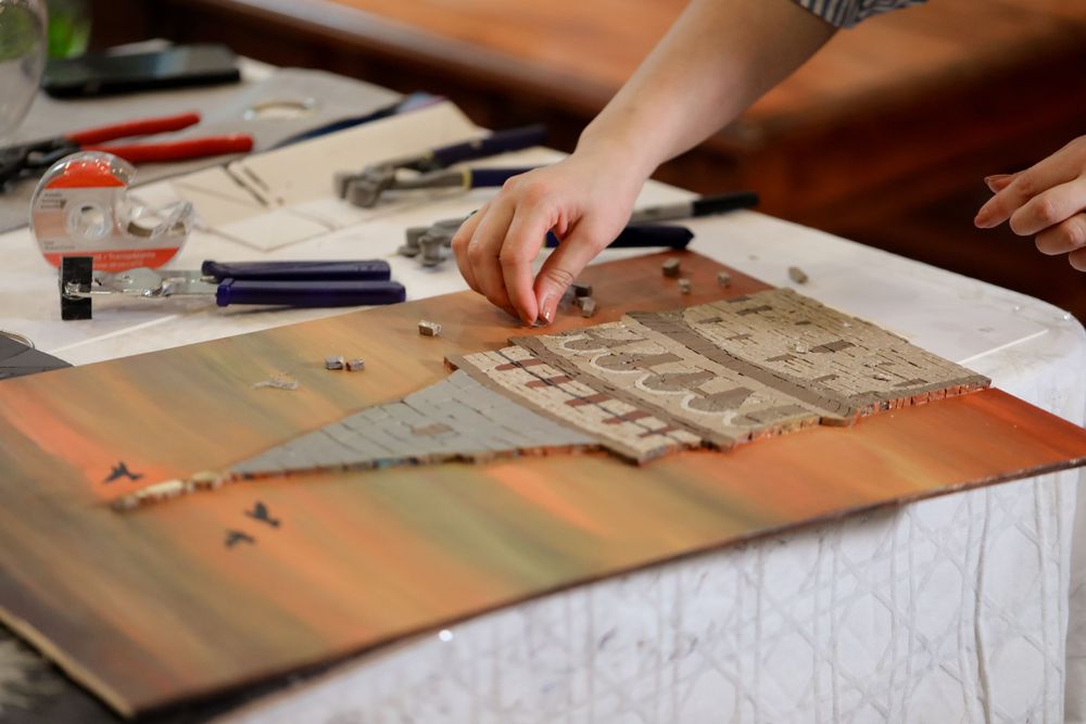 Hands working on a craft project with tools and paper materials spread across a wooden table surface.