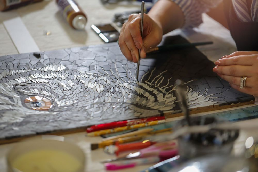 Artist's hands working on a detailed drawing with colored pencils and art supplies scattered on the workspace.