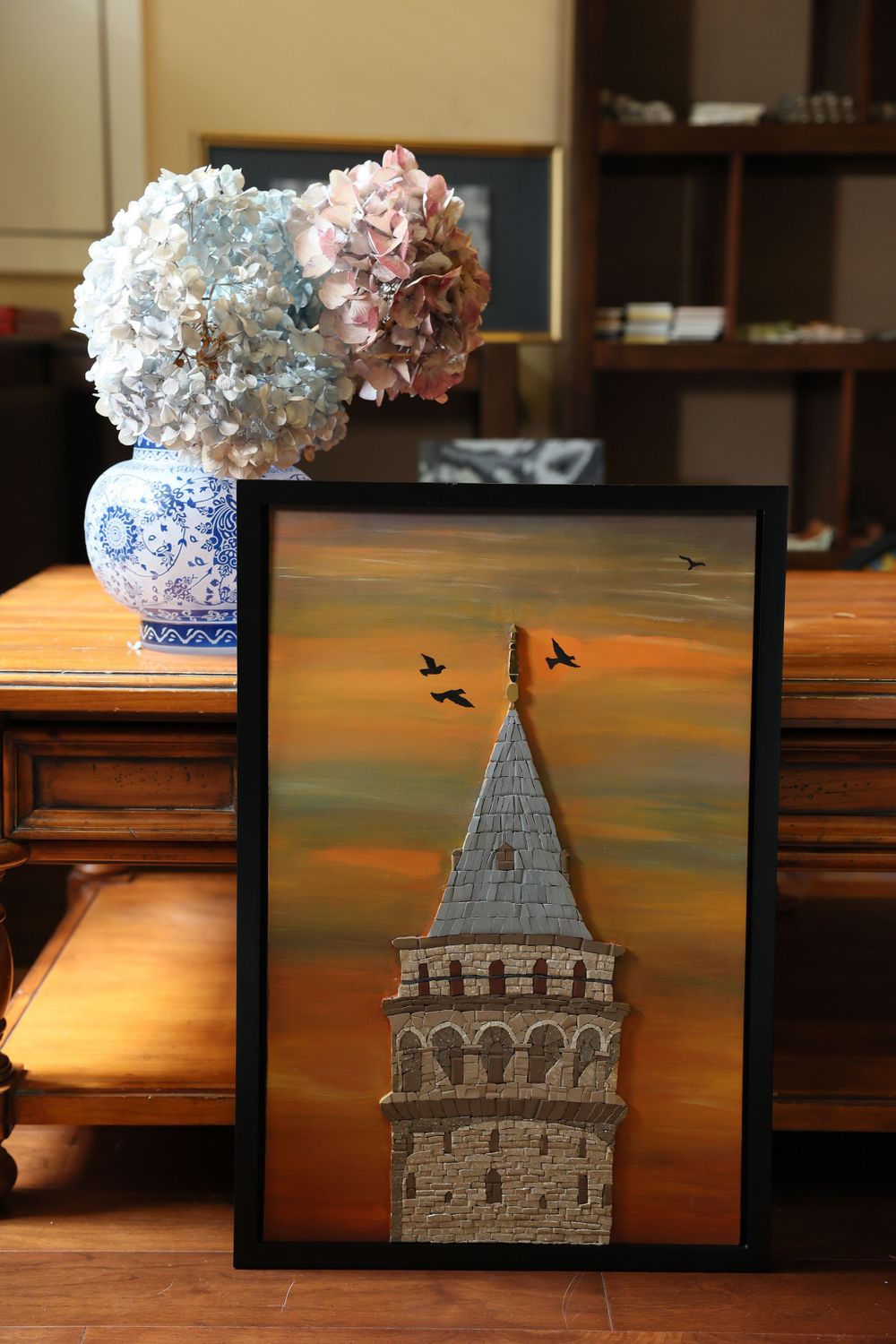 Framed artwork of Galata Tower against orange sunset sky with birds, next to blue and white vase with hydrangeas on wooden table.