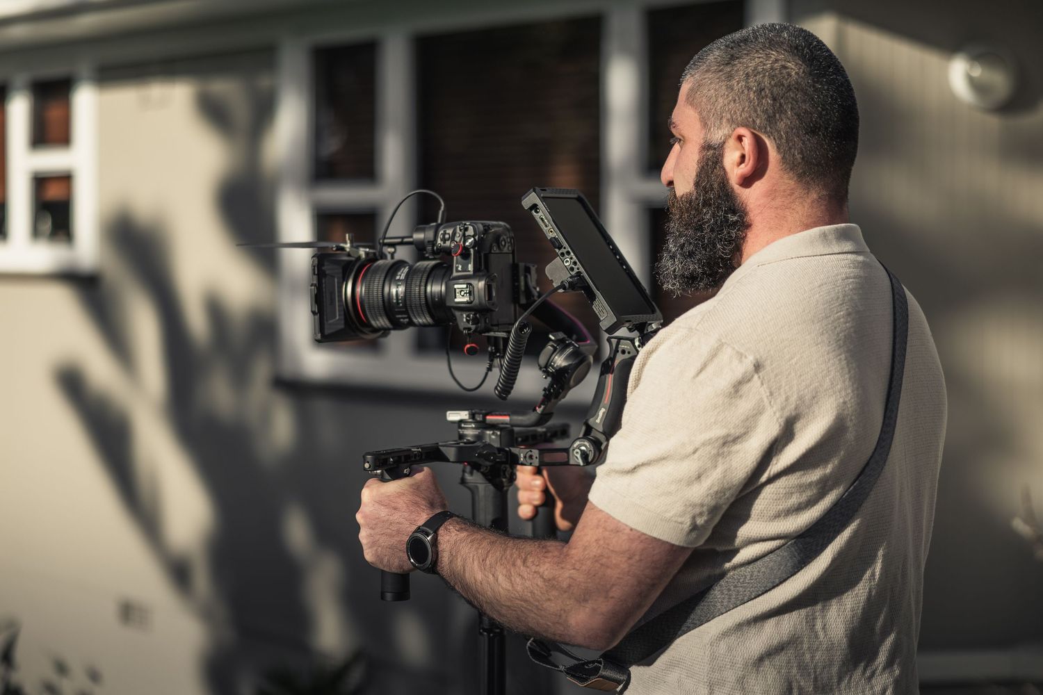 Professional camera operator with beard holds modern camera rig while filming outside in warm sunlight.