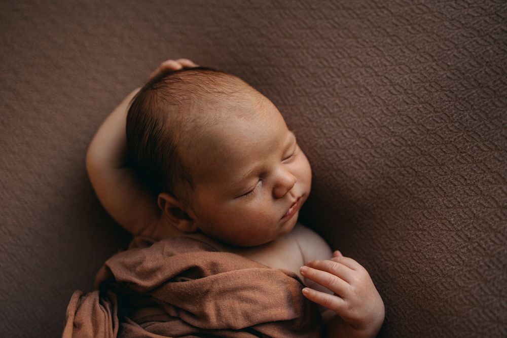 Peaceful sleeping newborn with delicate features rests on soft brown textured fabric in professional studio portrait.