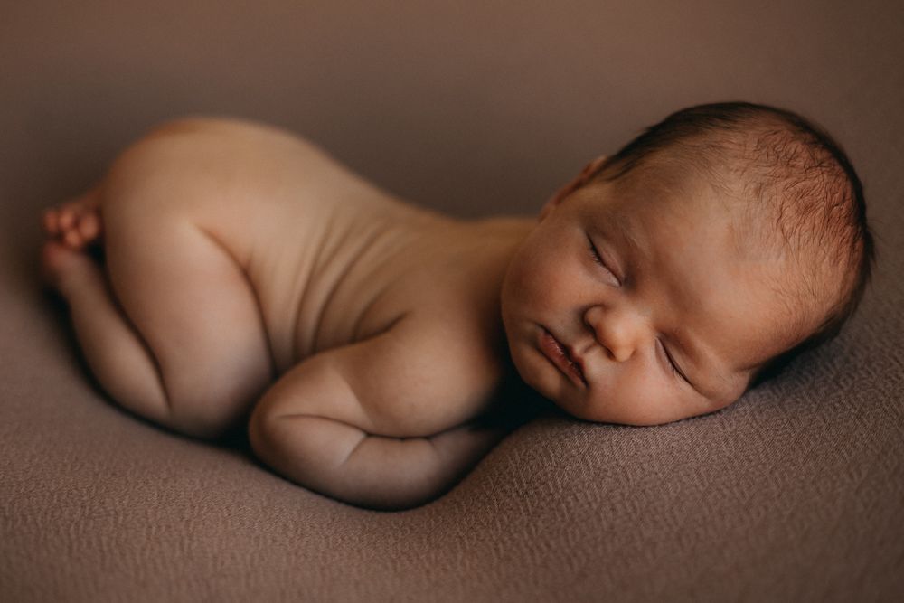 Artistic newborn photography of sleeping baby curled up on brown backdrop in natural pose.