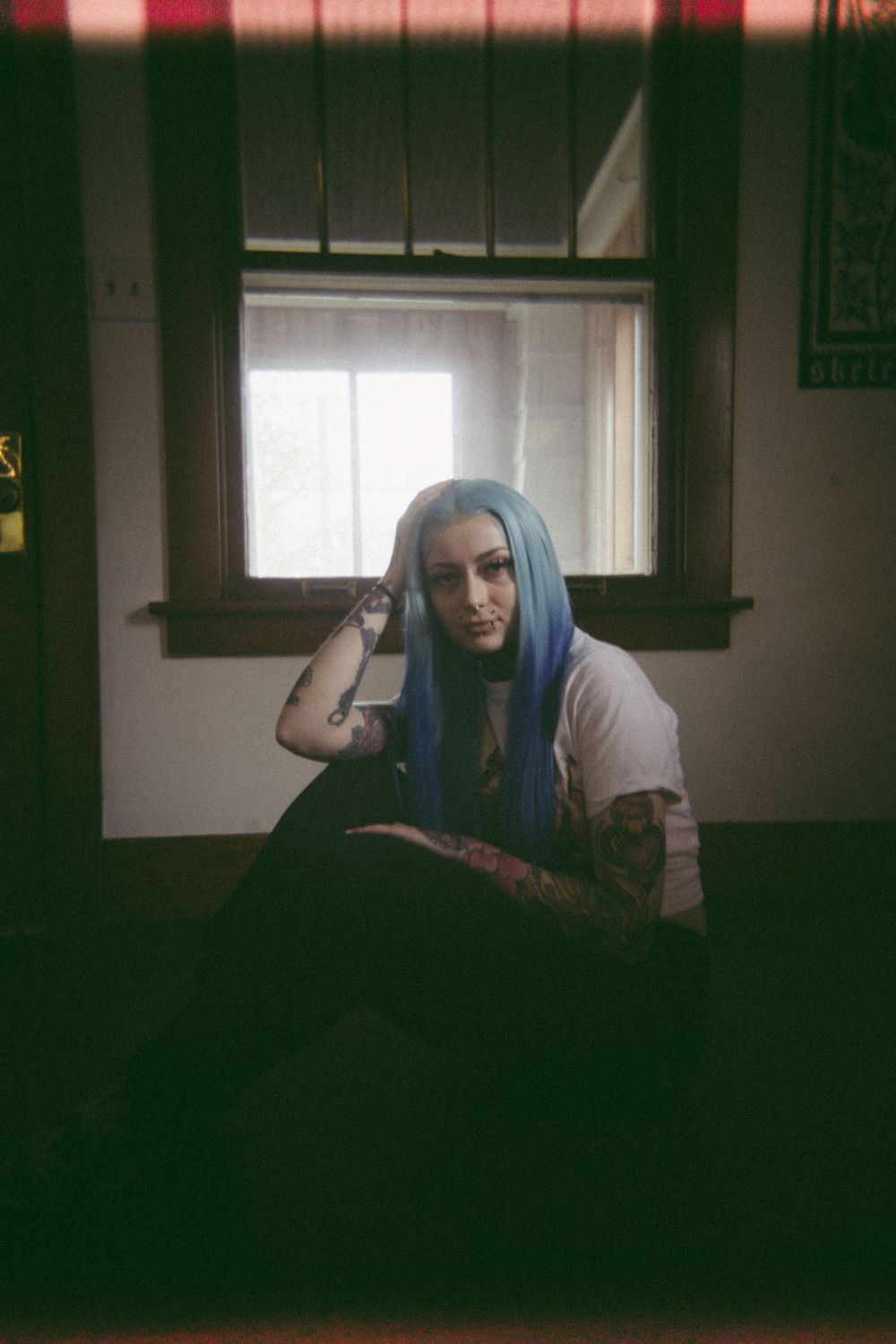 A dimly lit indoor scene shows someone with blue hair lounging near a window.