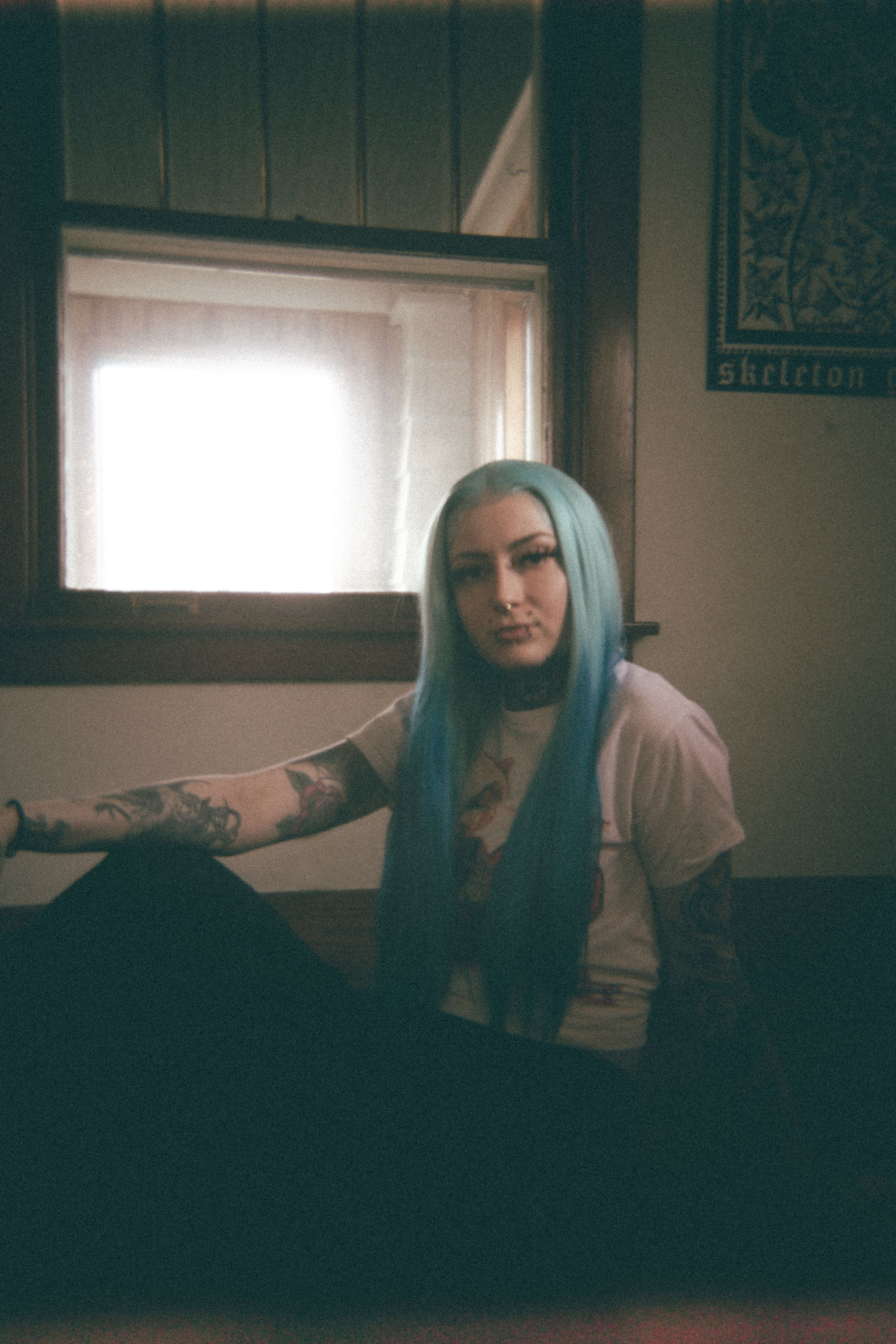 A moody indoor portrait features blue hair against a window-lit background.