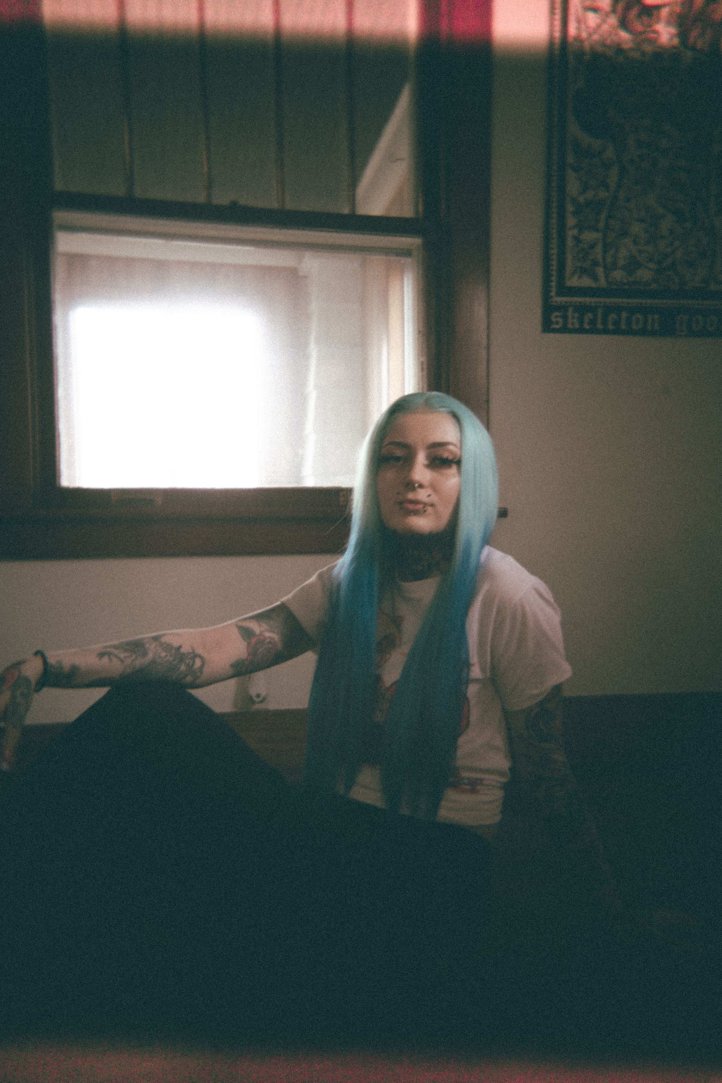 A person with blue hair poses in a moody indoor setting with natural window light.