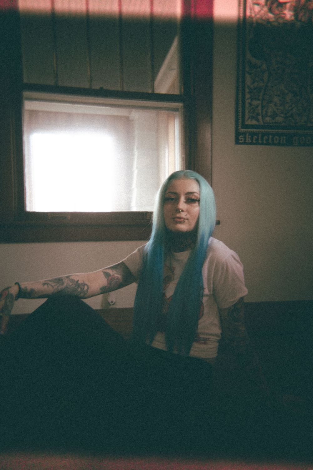 A person with blue hair poses in a moody indoor setting with natural window light.
