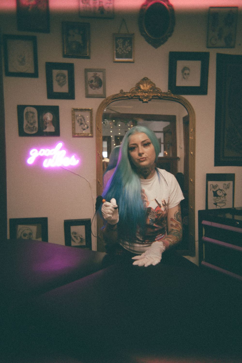 Indoor photoshoot with tattoos and blue hair against wall of vintage frames and neon signs.