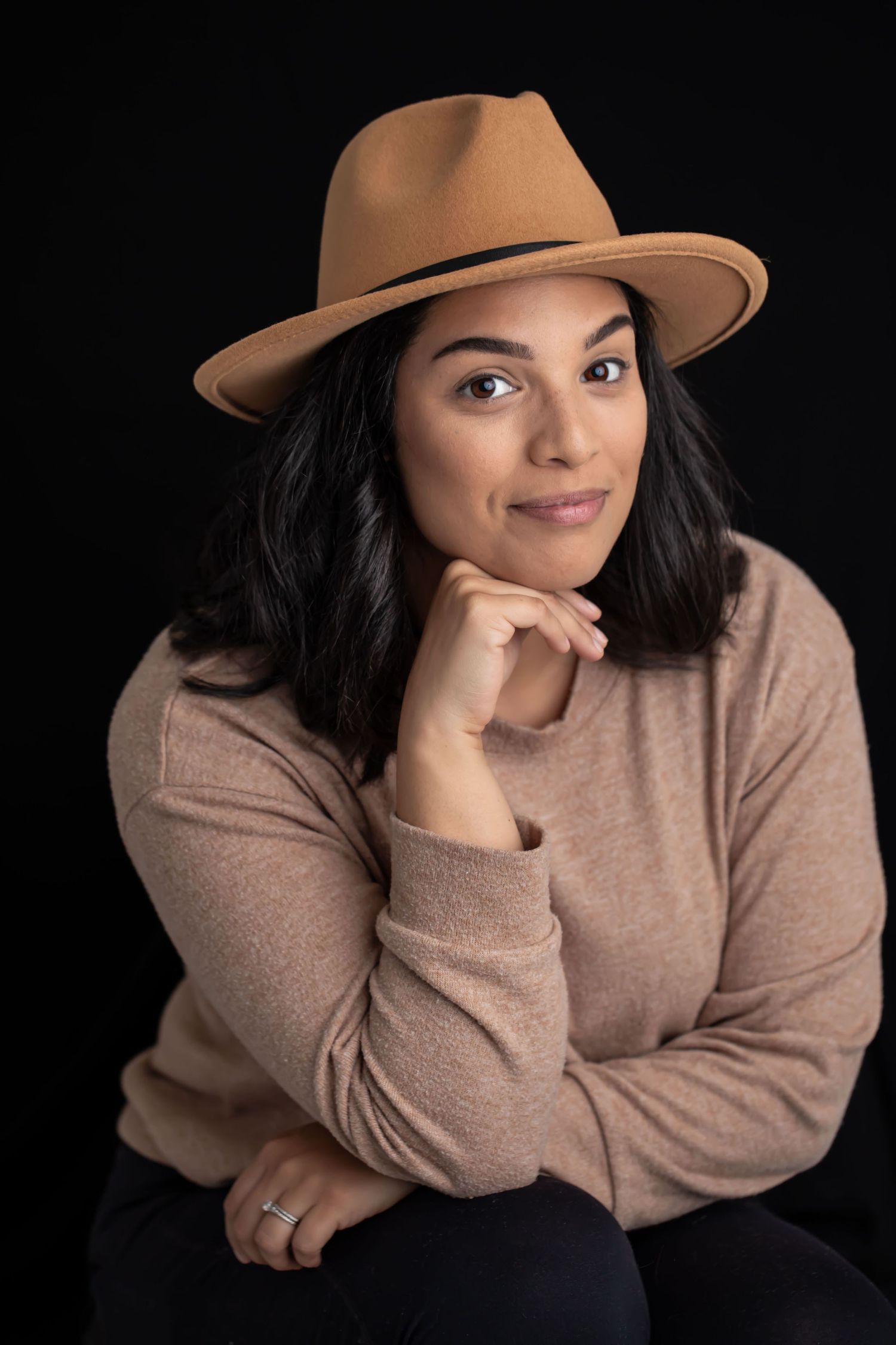 Portrait of a person wearing a tan fedora hat and beige sweater against a black background, resting chin on hand.