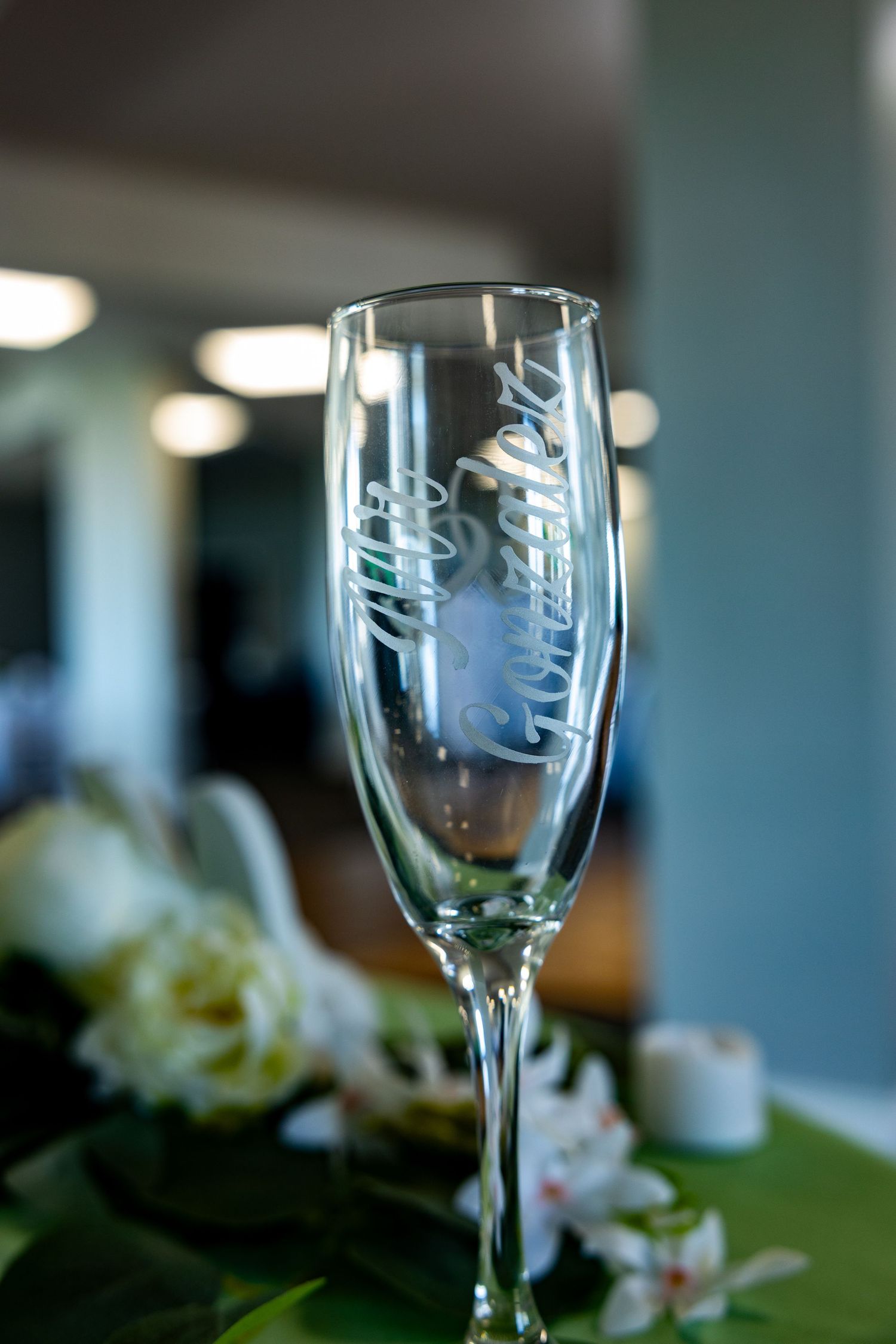 Elegant champagne flute with etched text sits on a green surface with blurred white flowers in background.