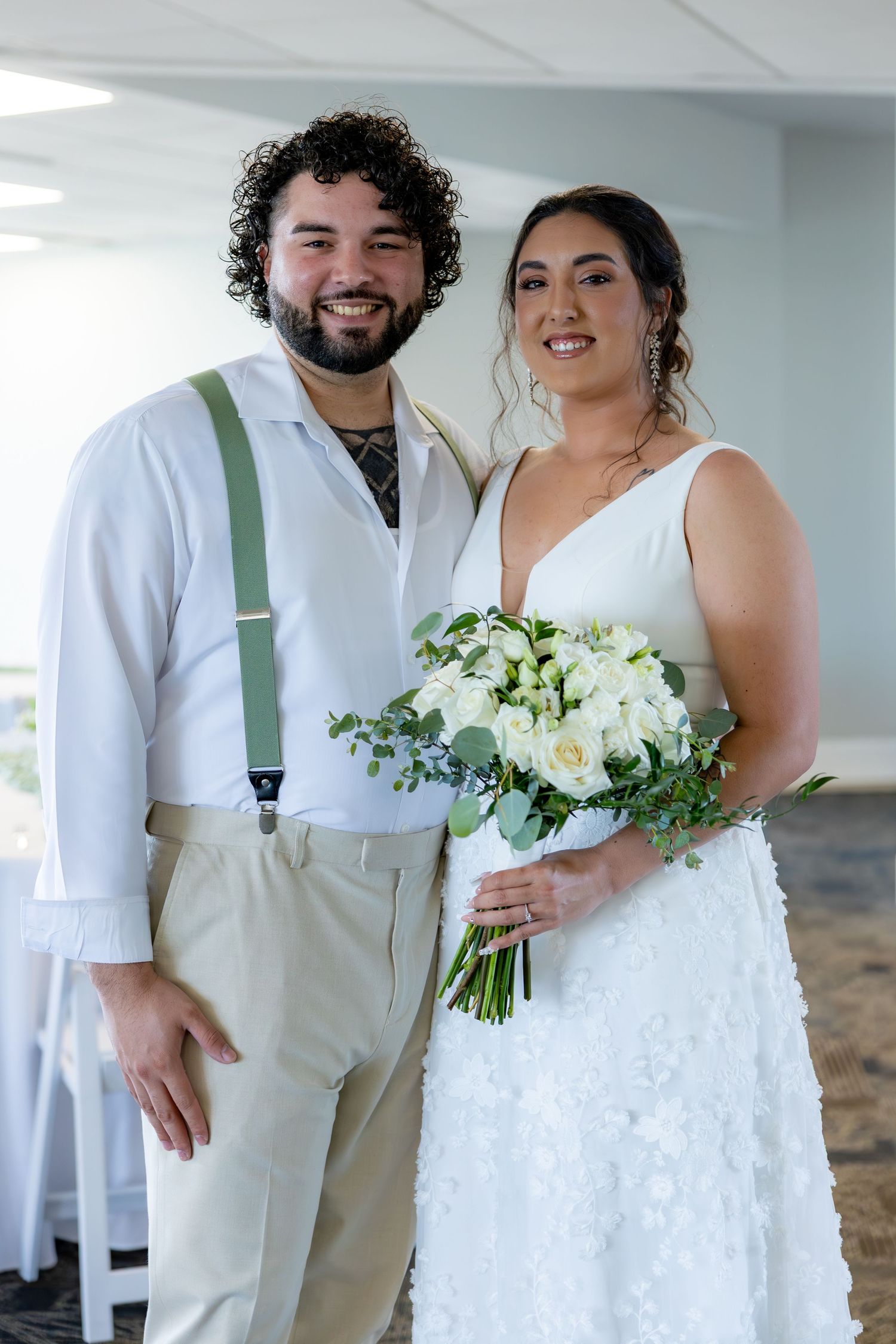 A couple pose together at their wedding, with the bride holding a white floral bouquet and the groom wearing suspenders.
