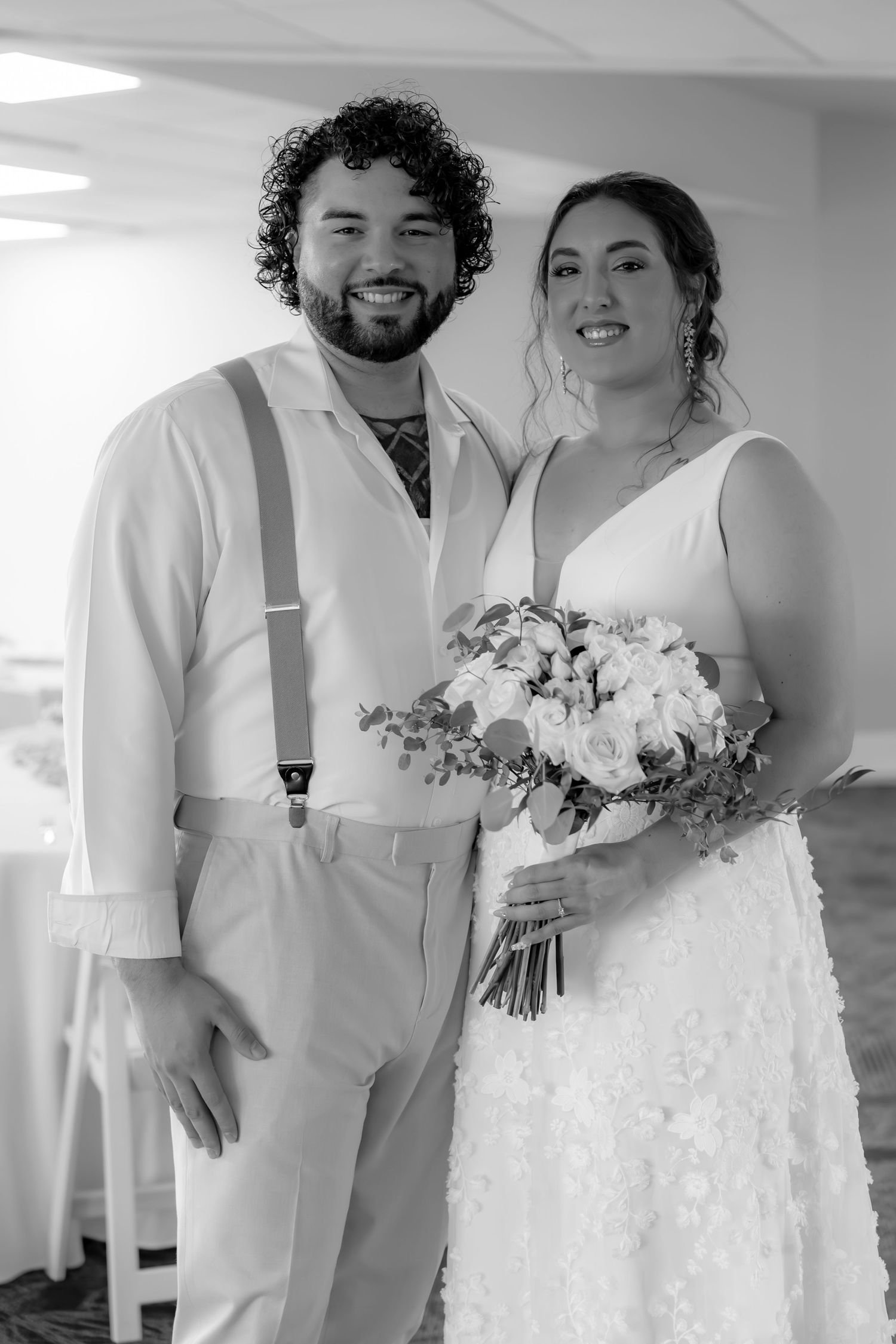 A bride in a white gown holds a bouquet while standing with her partner who wears a white suit with suspenders.