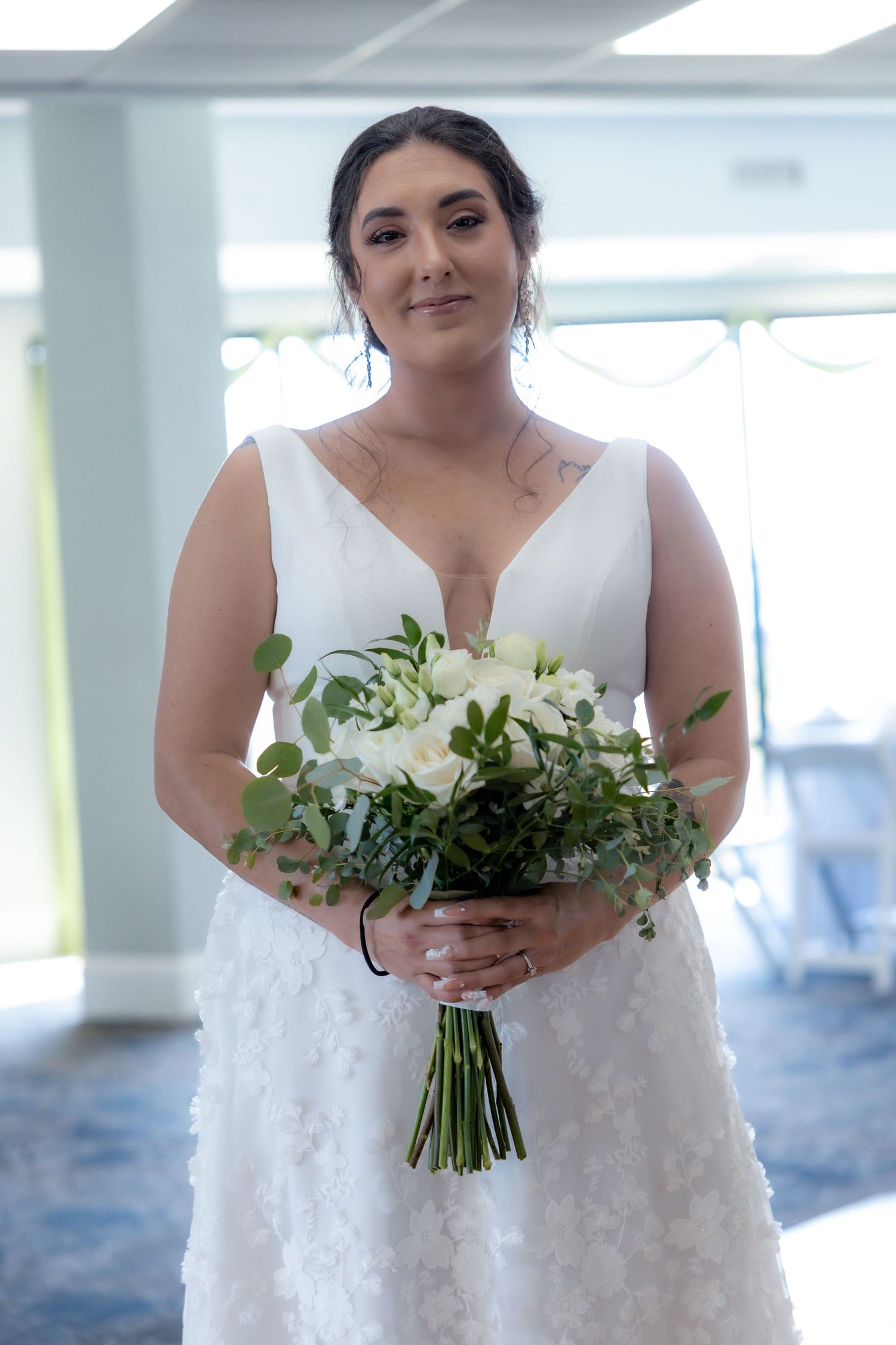 Beautiful bride in a sleeveless lace wedding dress holds an elegant white and green bouquet in a bright indoor space.