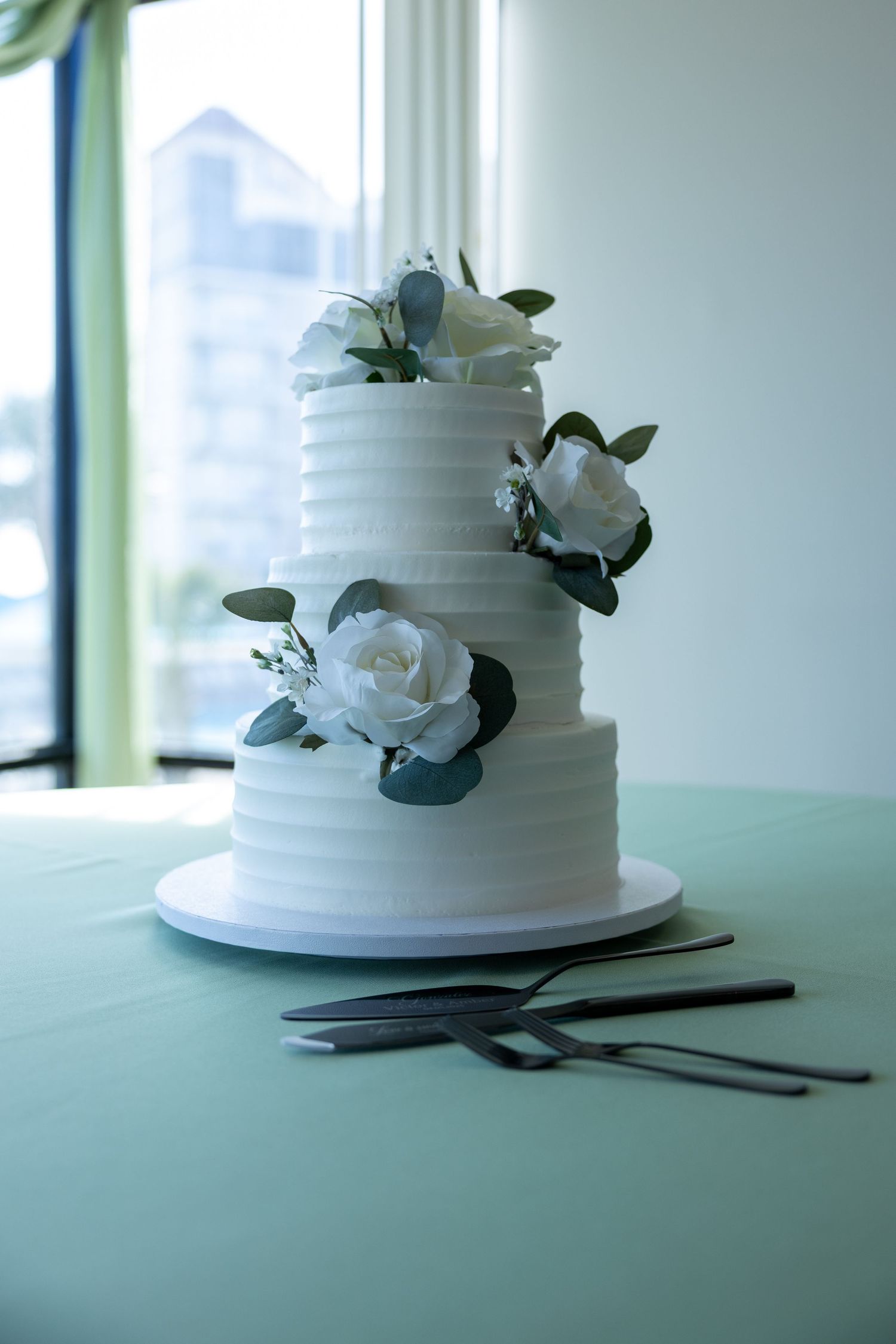 Elegant three-tier white wedding cake decorated with white roses and greenery sits beside serving utensils on turquoise table.