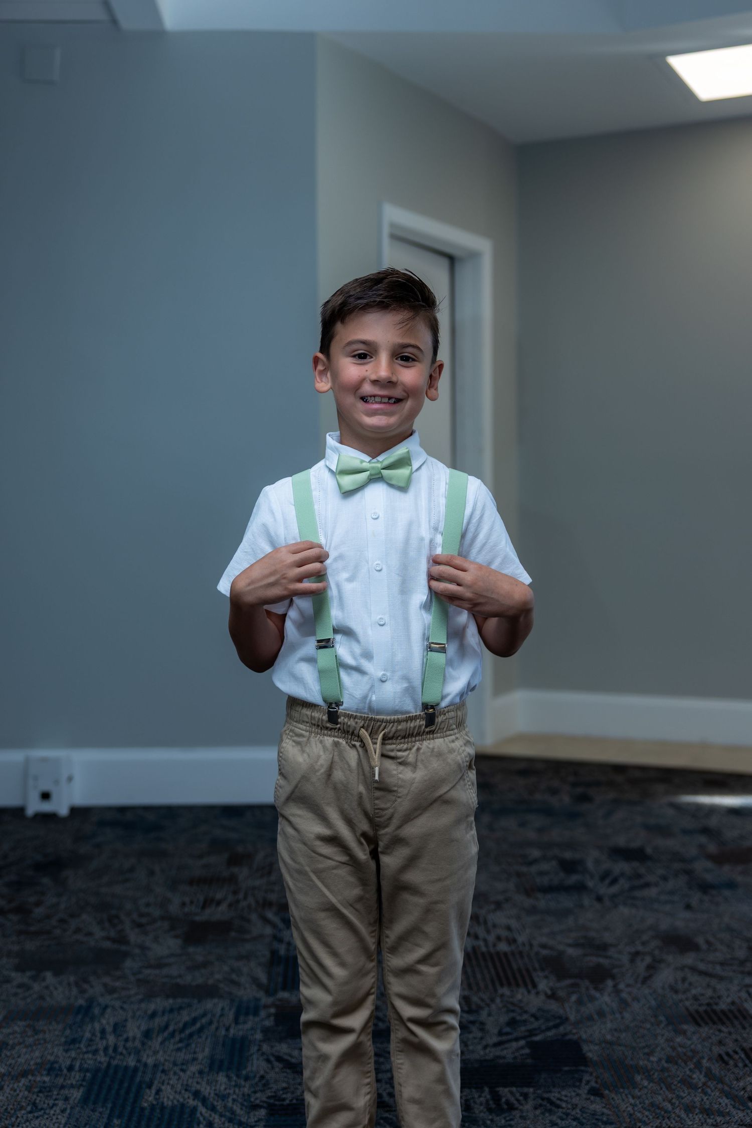 A young person in dress clothes wearing a mint green bow tie and suspenders standing in a room with gray walls.