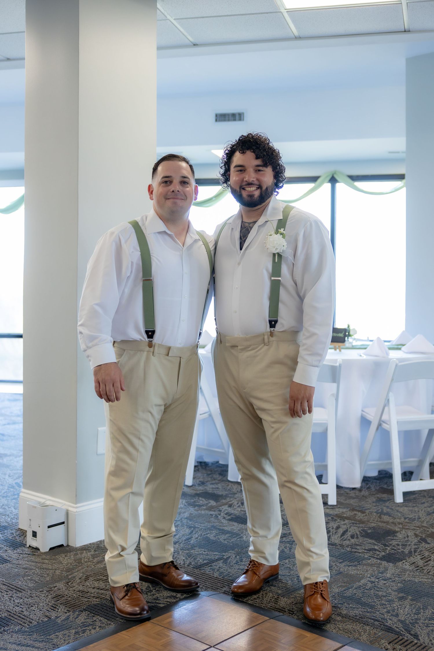 Two people pose together in matching light khaki pants, white dress shirts, and suspenders at a formal indoor event.