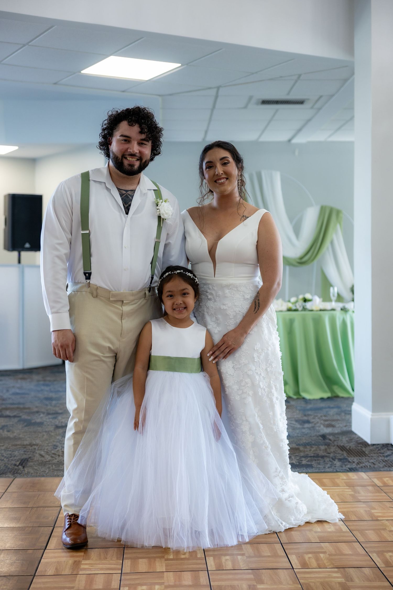 Wedding party members pose together on a wooden dance floor in a reception venue decorated with green accents.