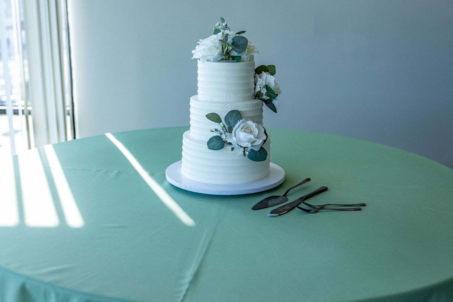 Three-tiered white wedding cake with delicate floral decorations sits on a mint green tablecloth in natural lighting.